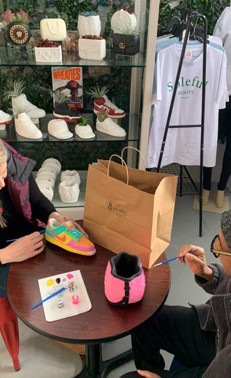 May include: A craft store display featuring painted sneaker-shaped planters and a variety of other decorative planters. A person is painting a pink planter. A colorful sneaker planter is on the table. A brown paper shopping bag is on the table.