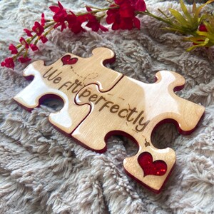 May include: Two wooden jigsaw pieces with red glitter hearts and the text "We Fit Perfectly" engraved on them. The pieces are on a light brown fluffy surface.