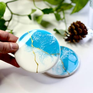May include: Two round coasters featuring a glossy finish with a white and bright blue abstract design. A thin, jagged gold line runs across the surface of each coaster, mimicking a crack pattern. The coasters have a smooth, reflective surface.