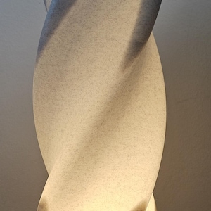 May include: A modern, sculptural table lamp with a twisted design. The lamp is made of a speckled, light-colored material, possibly ceramic or stone. The light shines through the form, creating shadows. The lamp is approximately 12 inches tall.