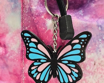 May include: A black butterfly charm with pink and blue wings hangs from a silver chain and a black cord loop. The charm is attached to a pink and purple galaxy-patterned backpack.
