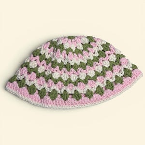 May include: A handmade crochet bucket hat in a granny square pattern. The hat features a color scheme of pink, white, and olive green. The brim and base are finished with a pink border. This hat is a stylish accessory.