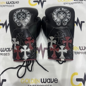 Puede incluir: Par de guantes de boxeo de cuero negro con costuras blancas y un diseño de cruz decorativa en rojo y blanco. Los guantes tienen el texto "ONE HEARTS" en un diseño circular blanco.
