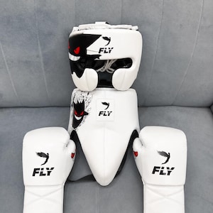 Op de afbeelding: Een witte boksset bestaande uit een hoofdbeschermer, een tok en twee bokshandschoenen. Elk onderdeel is voorzien van het zwarte FLY-logo en een afbeelding van een zwart wezen met rode ogen en een witte grijns op wit materiaal.