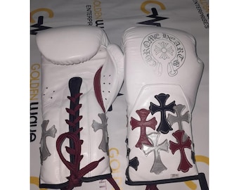 Könnte beinhalten: Weiße Leder-Boxhandschuhe mit dekorativen Kreuzen in Rot, Silber und Schwarz. Die Handschuhe haben eine Schnürung mit bordeauxfarbenen Schnürsenkeln und den Text "Chrome Hearts". Die Handschuhe sind für das Boxen konzipiert.