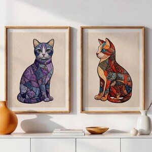 May include: Two framed wall art prints featuring stylised cats. The left print shows a cat with purple and blue mosaic-like patterns and a crescent moon collar. The right print displays a cat with orange, red, and blue geometric patterns.