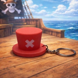 May include: A red top hat keychain with a white cross symbol, resting on a wooden surface. The background features a pirate ship and a seascape with a blue sky and clouds. The keychain is attached to a silver keyring.
