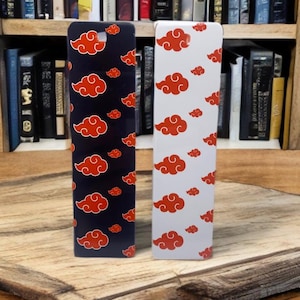 May include: Two phone cases, one black and one white, each adorned with a pattern of red cloud designs. The cases are rectangular and stand upright against a backdrop of a wooden surface and a bookshelf filled with books.