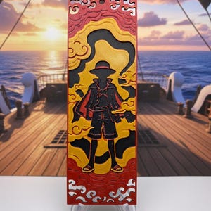 May include: A rectangular, layered artwork featuring a silhouette of a character in a hat and cape, set against a backdrop of yellow and red clouds. The artwork is displayed on a clear stand, with a ship and ocean scene in the background.
