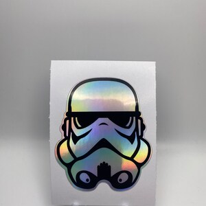 May include: A holographic sticker featuring a black and white illustration of a Stormtrooper helmet.