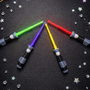 May include: Four toy lightsabers with green, purple, yellow, and red blades are arranged in a circle on a black surface. The hilts are black and gray. The background is decorated with stars and clear beads.