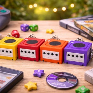 May include: A collection of colorful GameCube-shaped keychains in yellow, red, orange, and purple. The keychains have a black top and white front panel with three holes. Other gaming accessories, including game cartridges and a controller, are also visible.