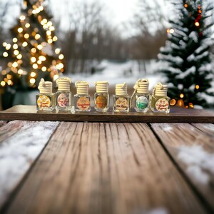 May include: A collection of small glass bottles with wooden caps, each labeled with a different scent name. The bottles are arranged on a wooden tray, with a snowy outdoor scene and Christmas trees in the background. The bottles are filled with a liquid.