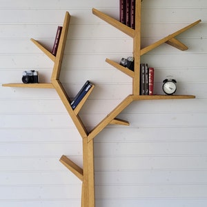 May include: A wooden bookshelf shaped like a tree with multiple branches and shelves. The shelves are filled with books, a camera, and an alarm clock.