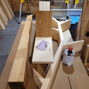 May include: A wooden shelf unit with a natural wood finish. The shelf unit is made of several pieces of wood that are joined together. There is a can of wood stain on the table next to the shelf unit.