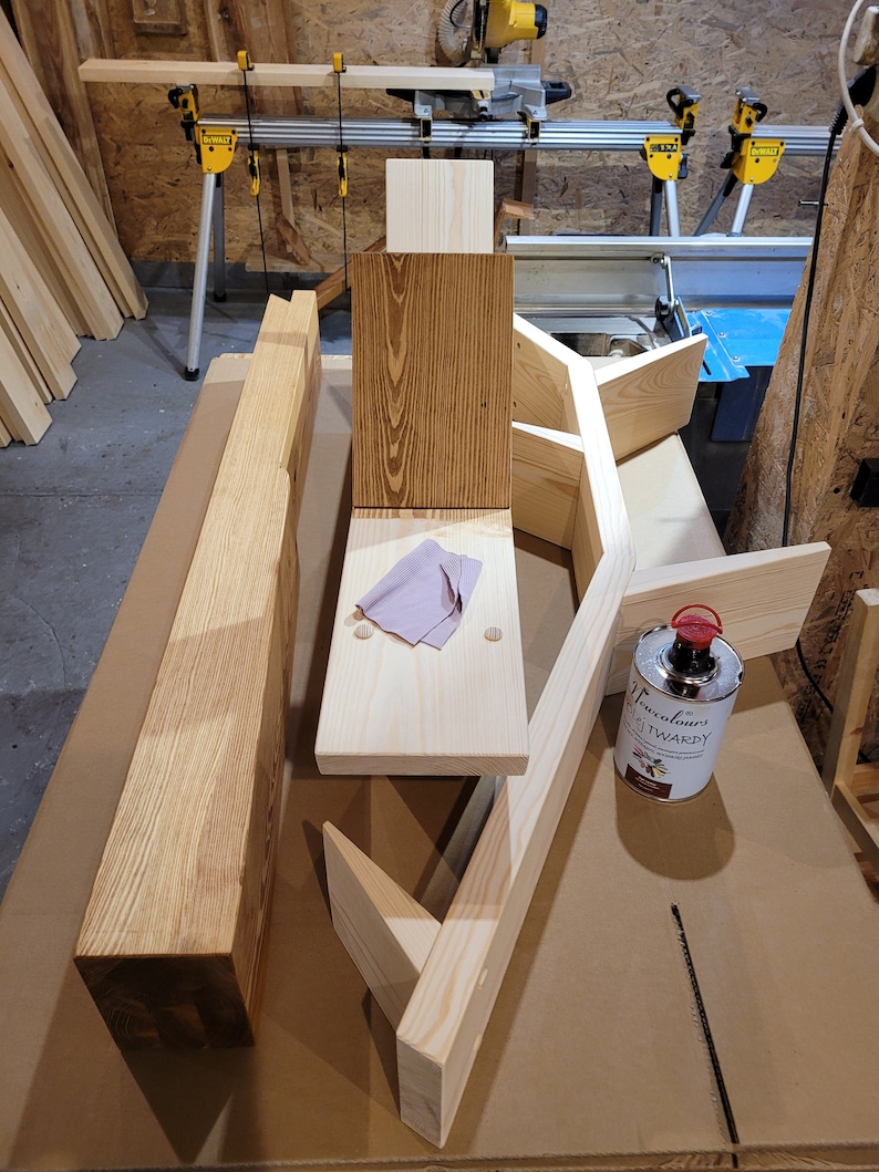 May include: A wooden shelf unit with a natural wood finish. The shelf unit is made of several pieces of wood that are joined together. There is a can of wood stain on the table next to the shelf unit.