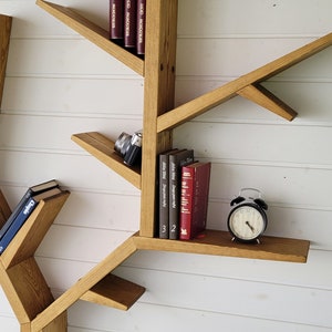 May include: A wooden tree-shaped bookshelf with multiple shelves and branches. The shelves are filled with books and a camera. A black alarm clock sits on one of the shelves.