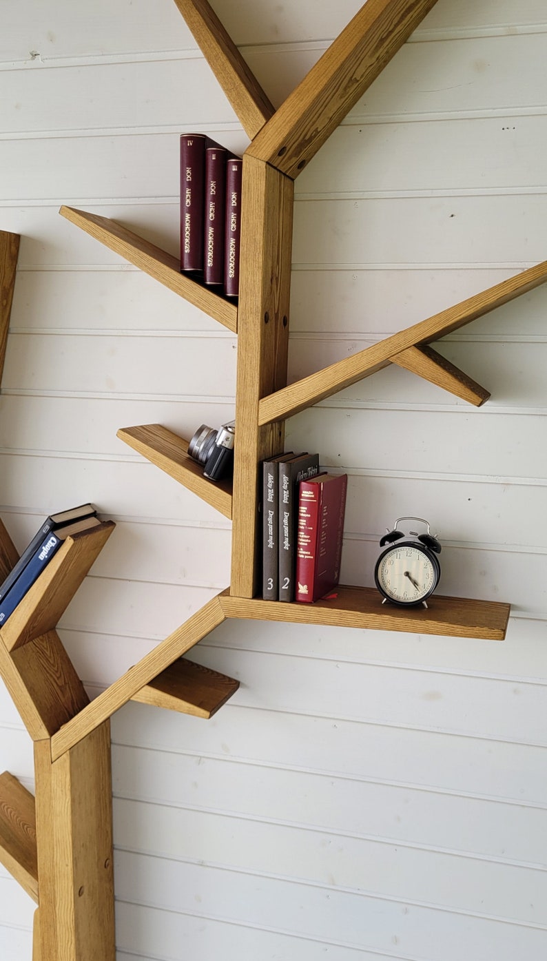 May include: A wooden tree-shaped bookshelf with multiple shelves and branches. The shelves are filled with books and a camera. A black alarm clock sits on one of the shelves.