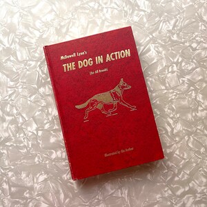 May include: A red book titled "THE DOG IN ACTION" by McDowell Lyon's. The cover features a gold illustration of a dog in motion. The text "(For All Breeds)" is also visible. The book is illustrated by the author.
