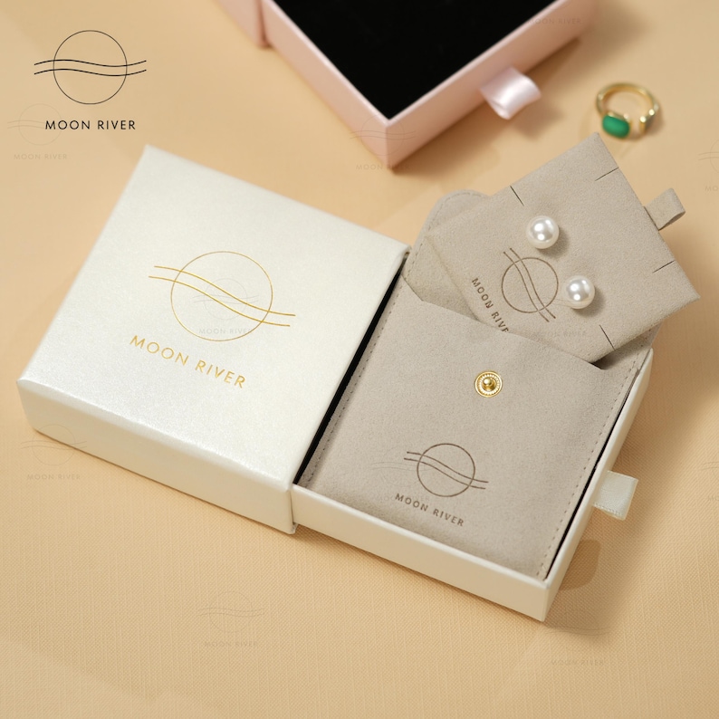 May include: A white box with a gold logo that says "Moon River" is open to reveal a smaller beige box with a gold logo that says "Moon River". The smaller box has a snap closure and contains a small white pouch with two pearl earrings.