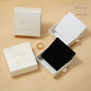 May include: Three white boxes with a gold logo that says "Moon River" and a wavy line design. The boxes are for jewelry and have a black velvet interior. One box is open and a gold ring is sitting on top of the black velvet.