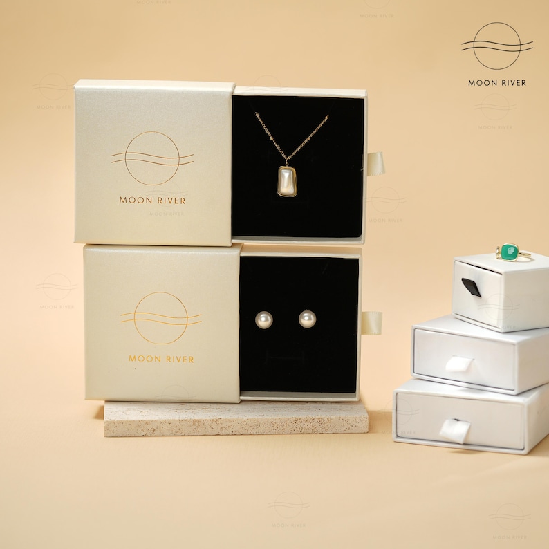 May include: Three white boxes with black interiors, each containing jewelry. The top box contains a gold necklace with a pearl pendant. The middle box contains a pair of pearl earrings. The bottom box is empty. All boxes have the logo "Moon River" printed on the front.