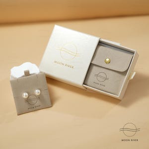 May include: A white box with a gold logo that says "Moon River" and a smaller beige box with a gold button and the same logo inside. The smaller box contains two pearl earrings.