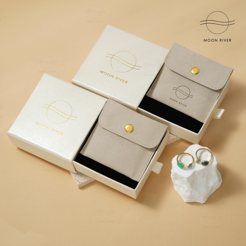 May include: Three white gift boxes with a gold logo that says "Moon River" and a beige velvet pouch inside. The boxes are open and the pouch is partially pulled out. Two rings are on a white rock next to the boxes.
