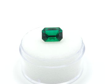 May include: A vibrant green emerald gemstone in a rectangular cut, displayed in a clear plastic container with a white foam interior.