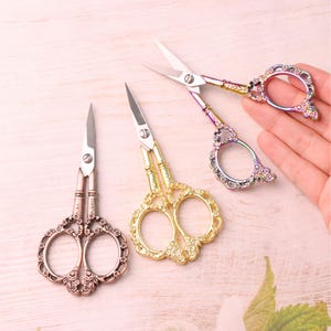 May include: Three pairs of metal embroidery scissors with ornate, floral-patterned handles. One pair is finished in a copper tone, one in a bright gold tone, and one features a multi-colored iridescent finish with sharp, pointed steel blades.