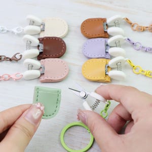 May include: A selection of small, colourful leather pouches with metal clasps and chains. The pouches are in pastel shades like cream, brown, lavender, and yellow. A pair of small scissors and a green ring are also in the image.