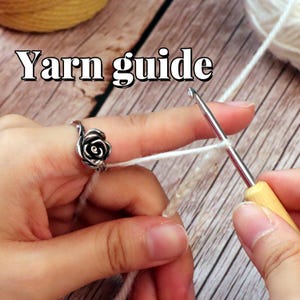 Könnte beinhalten: Nahaufnahme von Händen, die mit weißem Garn und einer Häkelnadel arbeiten. Ein silberner Rosenring befindet sich am Zeigefinger. Der Text "Yarn guide" wird in weißer Schrift mit schwarzer Umrandung angezeigt.