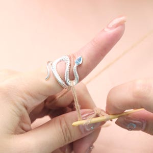 May include: A silver snake ring with a blue gemstone accent is worn on a finger. The ring is wrapped around the finger, and the snake's head is at the tip. The image shows a person crocheting with a wooden hook and yarn.