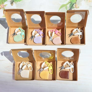 May include: A selection of small nail scissors in various colours, each paired with a matching leatherette sheath, presented in individual cardboard boxes with clear lids. The scissors have white handles and colourful accents.