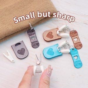 Può includere: Una collezione di piccoli e colorati accessori a tema filato, tra cui forbici, segnapunti e tiretti per cerniere. Gli accessori presentano disegni a cuore e filato in vari colori come grigio, beige e blu. Il testo "Small but sharp" è visualizzato.