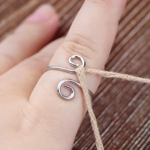 May include: A silver-colored yarn guide ring with a swirling design is worn on a finger, with a strand of beige yarn threaded through it. The ring is made of a thin, shiny metal.