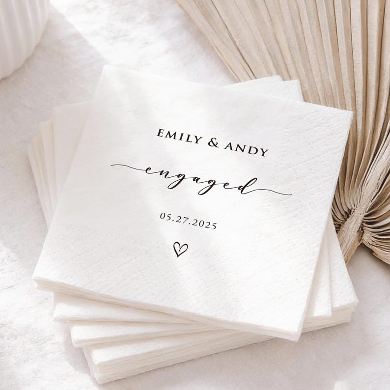 May include: A stack of white paper napkins with black text. The top napkin reads "EMILY & ANDY engaged 05.27.2025" with a small heart symbol. The napkins are stacked neatly, suggesting a wedding or engagement celebration.