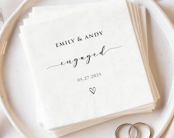 May include: A pile of white paper napkins printed with "EMILY & ANDY engaged 05.27.2025" in black script, with a heart symbol. Two silver wedding rings are on a white plate, perfect for an engagement.