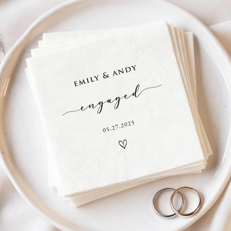 May include: A stack of white paper napkins with the words "EMILY & ANDY engaged 05.27.2025" printed in black script. A small heart is below the date. Two silver wedding rings are on a white plate.