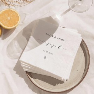May include: A stack of white paper napkins with the words "EMILY & ANDY engaged 05.27.2025" printed in black script. A small heart is printed below the date. The napkins are on a beige plate, with a lemon half visible.