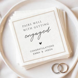以下が含まれることがあります： 金色の縁取りと「Pairs Well With Getting Engaged」の文字が入った白い紙ナプキンの束。その下に小さな黒いハートと「Emma & Jason」の名前があります。2つの指輪が前景にあります。