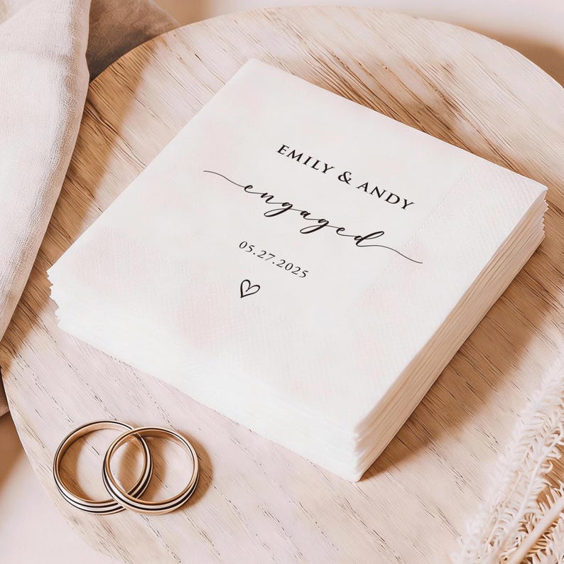 May include: A stack of white paper napkins with the words "EMILY & ANDY engaged" printed in black script, along with the date 05.27.2025 and a heart symbol. Two gold wedding rings are in the foreground.