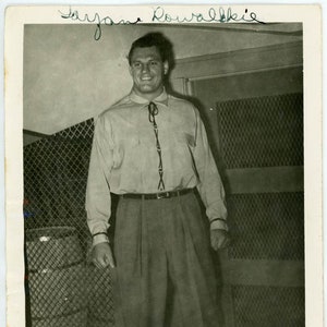 Puede incluir: Una fotografía en blanco y negro de un hombre que lleva una camisa de color claro con una corbata oscura y pantalones oscuros. El hombre sonríe y mira a la cámara. La foto está firmada "Wayne Roubalkie".