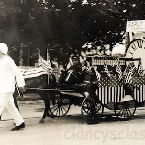 May include: Vintage black and white photo of a parade float pulled by a horse. The float features a large clock, American flags, and the text "Early Birds Committee". A person in white attire walks alongside the horse.
