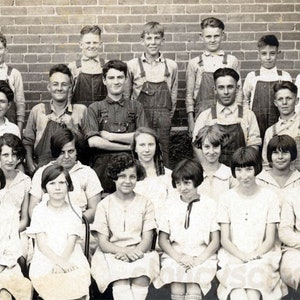 May include: A black and white photo of a class of students, mostly wearing overalls, posing for a photo in front of a brick wall.