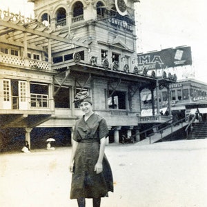May include: A vintage black and white photograph showing a person wearing a dark short-sleeved dress, a patterned headscarf, and dark leggings with lace-up shoes, standing in front of a large multi-story building labeled Convention Hall.