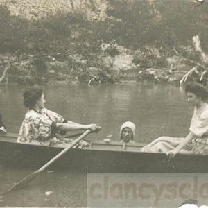 May include: Vintage black and white photograph of a group of people in a rowboat on a body of water. One person is rowing, while others are seated. The background shows trees and foliage.