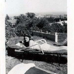 May include: A black and white photo of a woman wearing a hat, lying on a lounge chair in a garden. The woman is looking at the camera. The photo was taken in the 1950s.