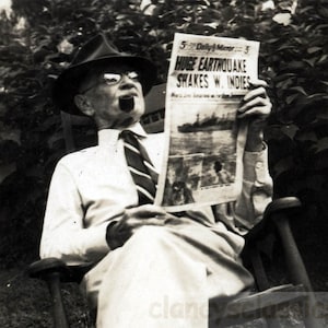 May include: Black and white photograph of a man in a hat and glasses smoking a pipe while reading a newspaper. The newspaper headline reads "Huge Earthquake Shakes W. Indies."