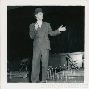 May include: A black and white photo of a man in a suit and fedora, standing on a stage with a microphone in his hand. He is gesturing with his right hand. There are chairs and a white fence in the background.